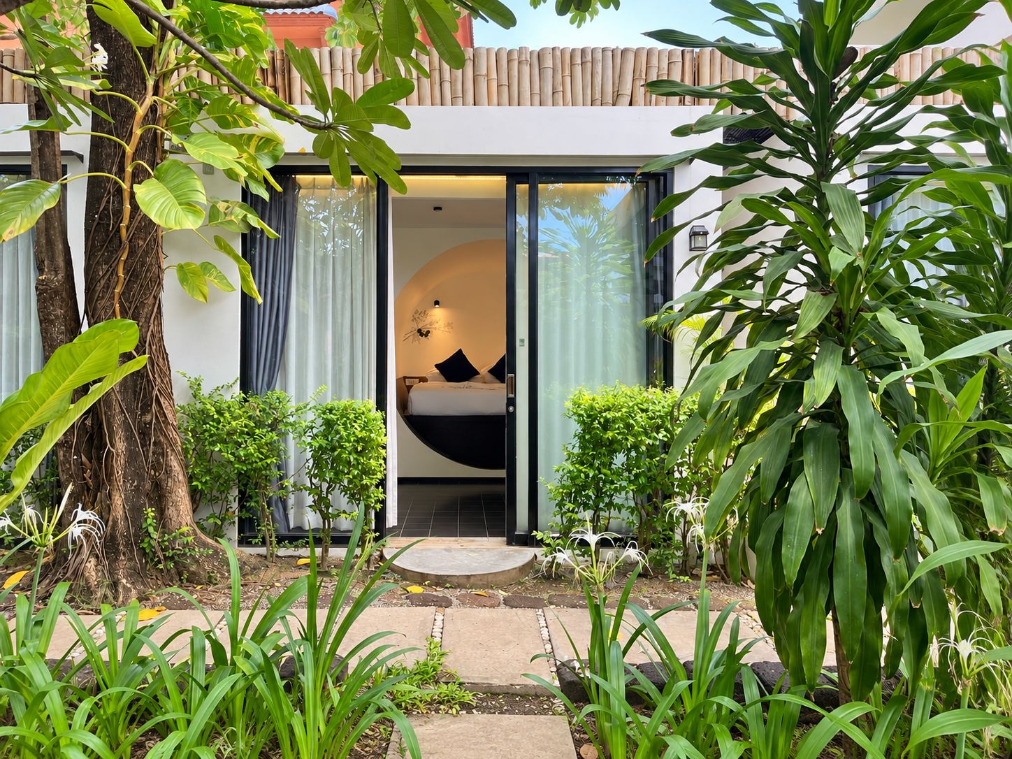 A modern building with a glass door entrance, a bed with black bedding, and a small garden area with various plants.