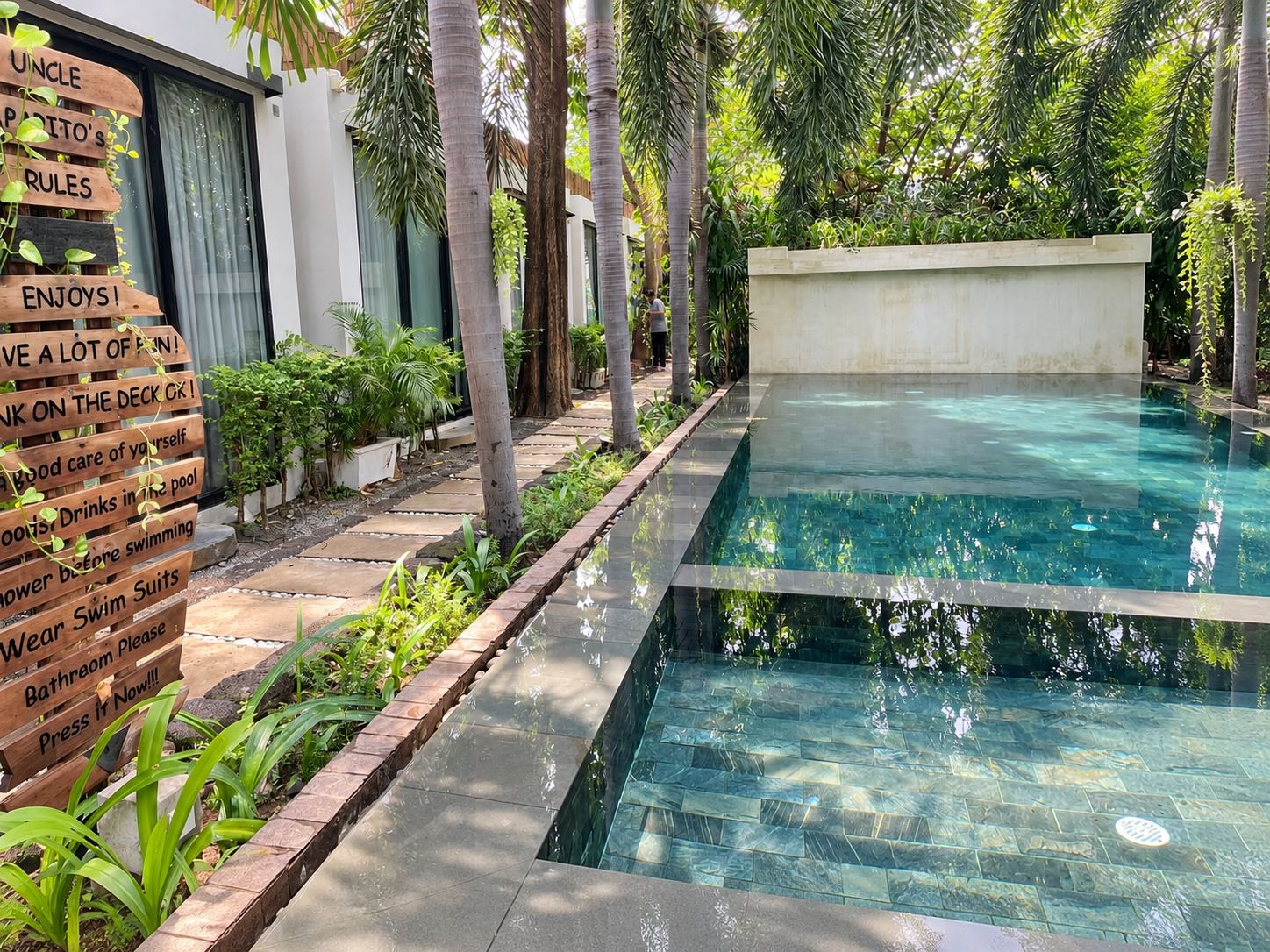Piscina con un letrero de madera que enumera normas, rodeada de vegetación y un edificio moderno.