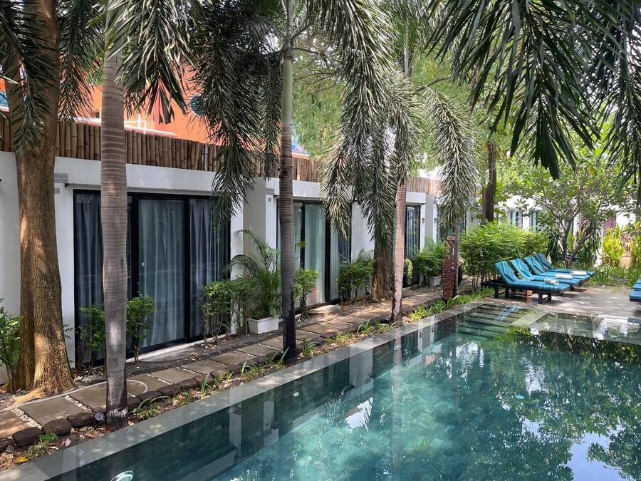 Un área de piscina con sillones reclinables, palmeras y un edificio con amplias ventanas.