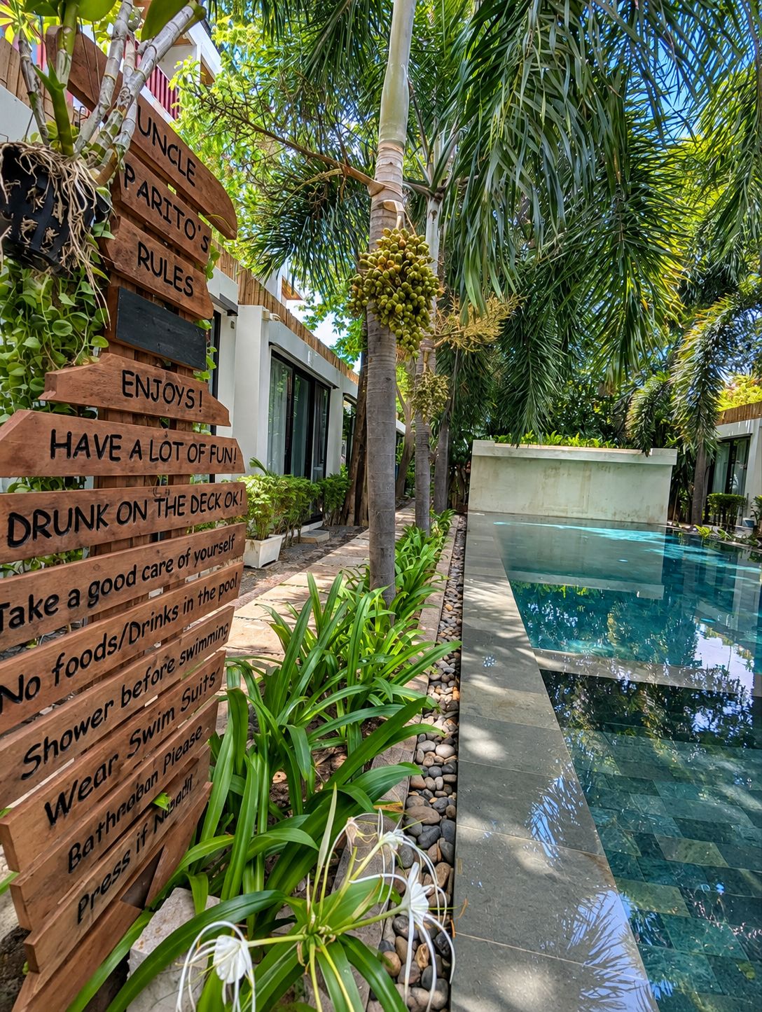 Señal de madera con diversas normas para la zona de la piscina, rodeada de exuberante vegetación y una piscina cristalina.