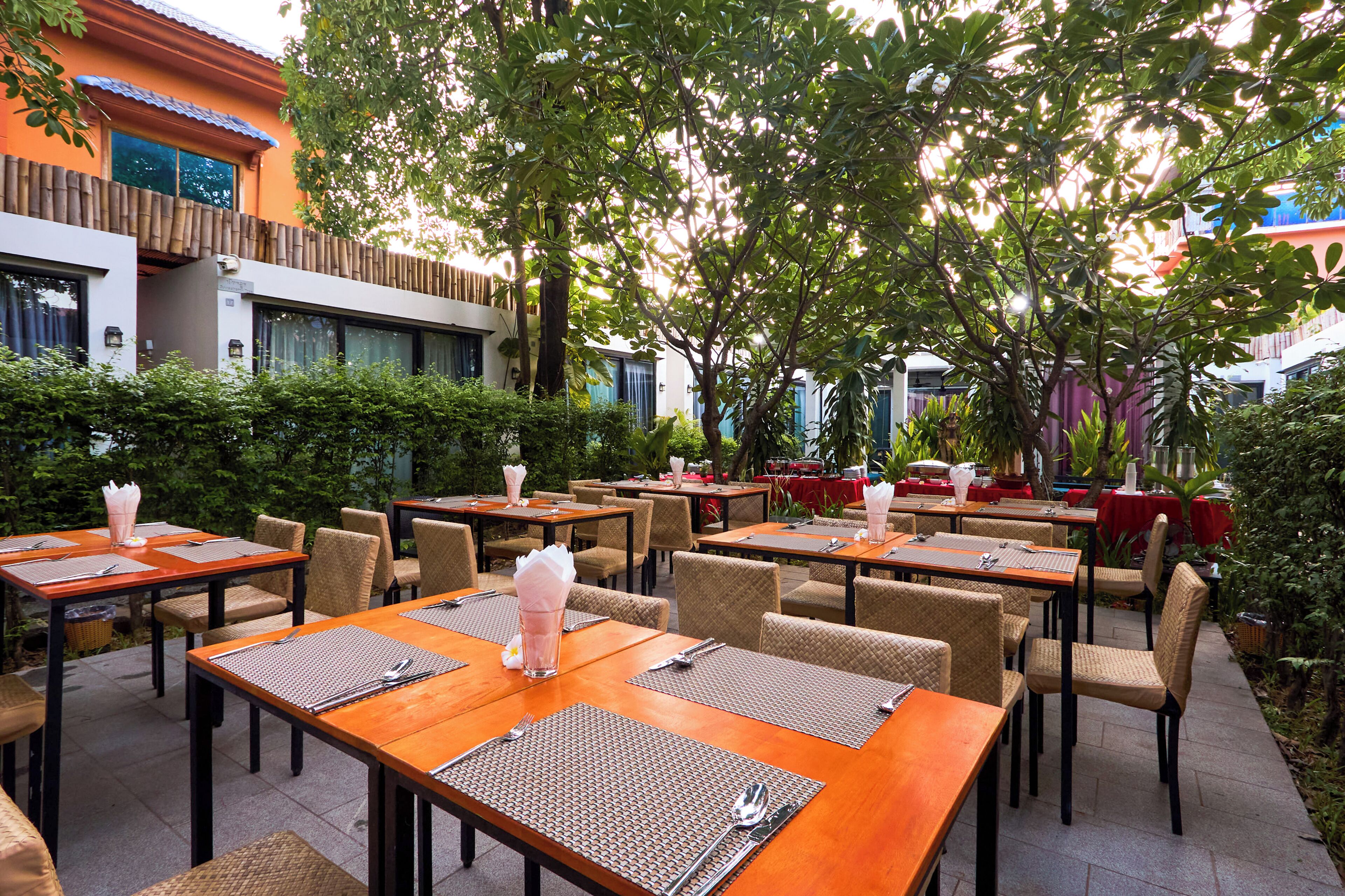 An outdoor dining area with tables and chairs, surrounded by greenery and buildings.