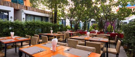 An outdoor dining area with tables and chairs, surrounded by greenery and buildings.