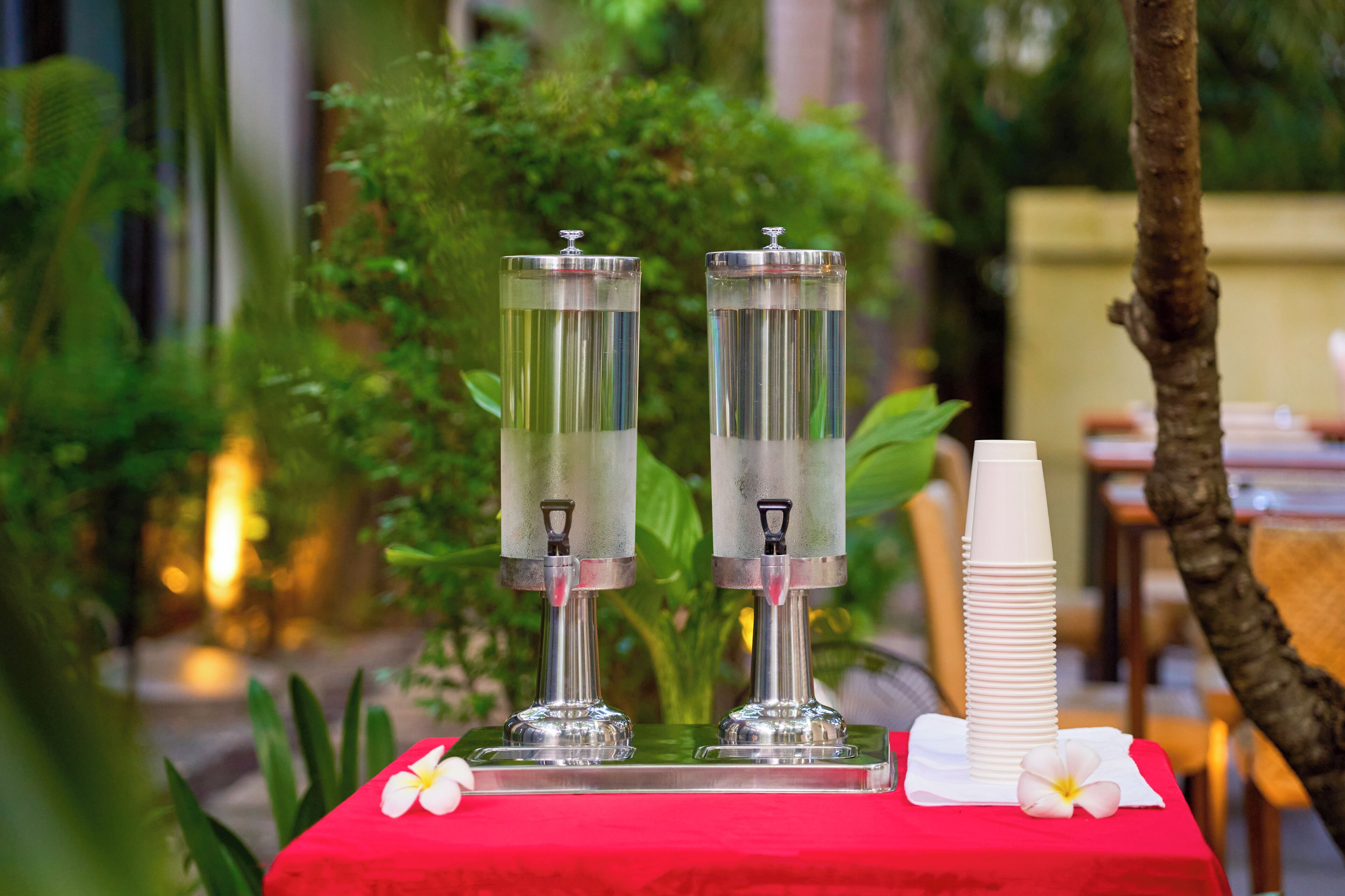 A table with two metallic water dispensers, a stack of white paper cups, and a red tablecloth.