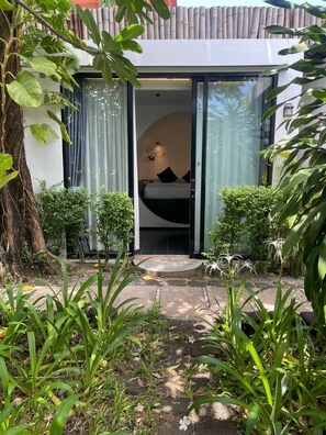 Un bâtiment moderne avec une entrée dotée d’une porte en verre, un petit jardin agrémenté de plantes et d’un arbre, et un lit visible à l’intérieur.