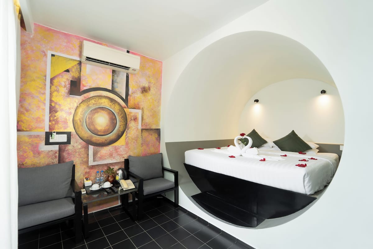 Quarto de hotel moderno com teto curvo singular, uma cama grande com pétalas de rosa, um sofá cinzento, uma mesinha com um telefone e uma garrafa, e uma pintura abstrata colorida na parede.