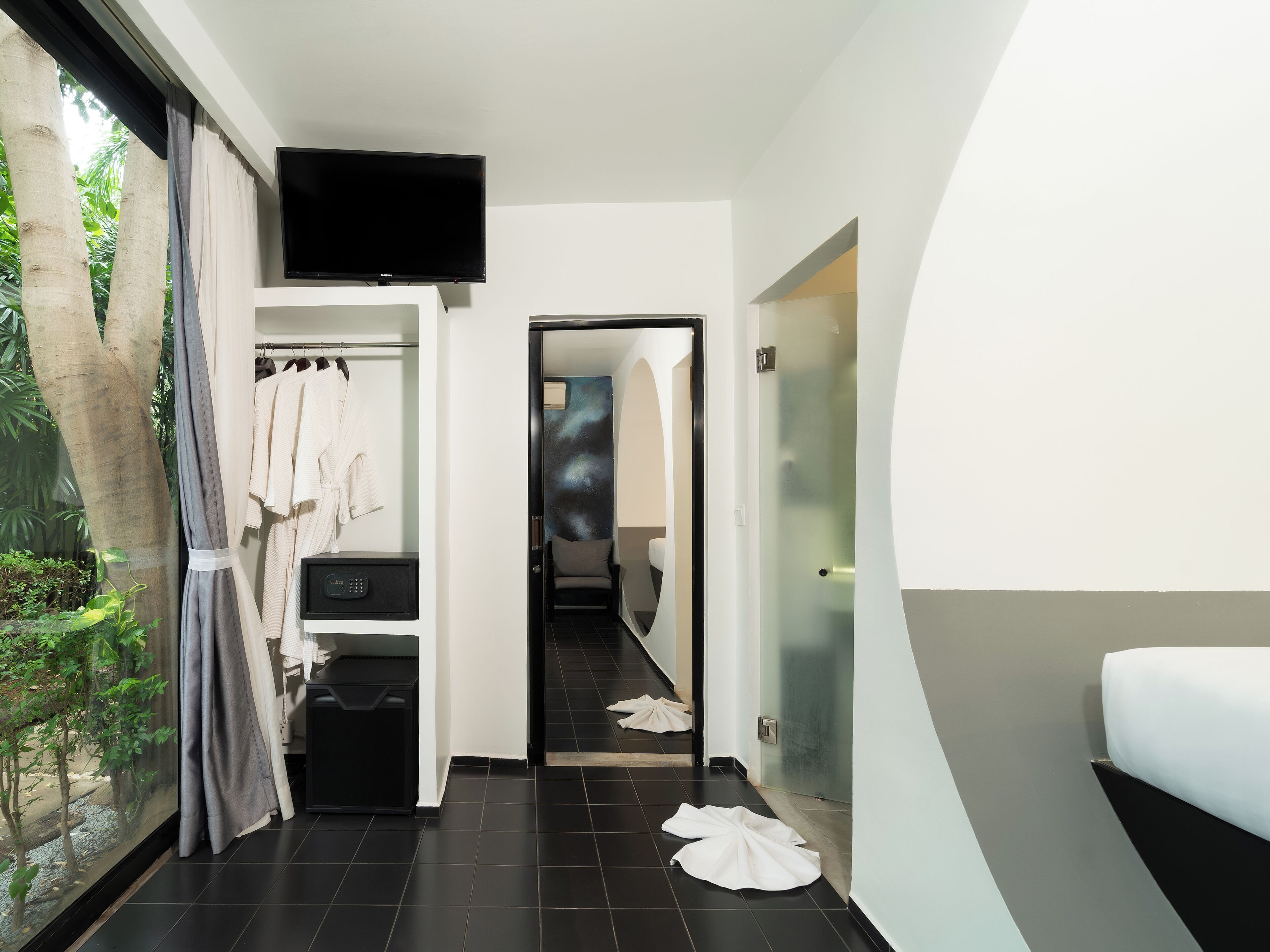 A modern hotel room with a large window, a flat-screen TV, a walk-in closet, and a bathroom with a glass door.
