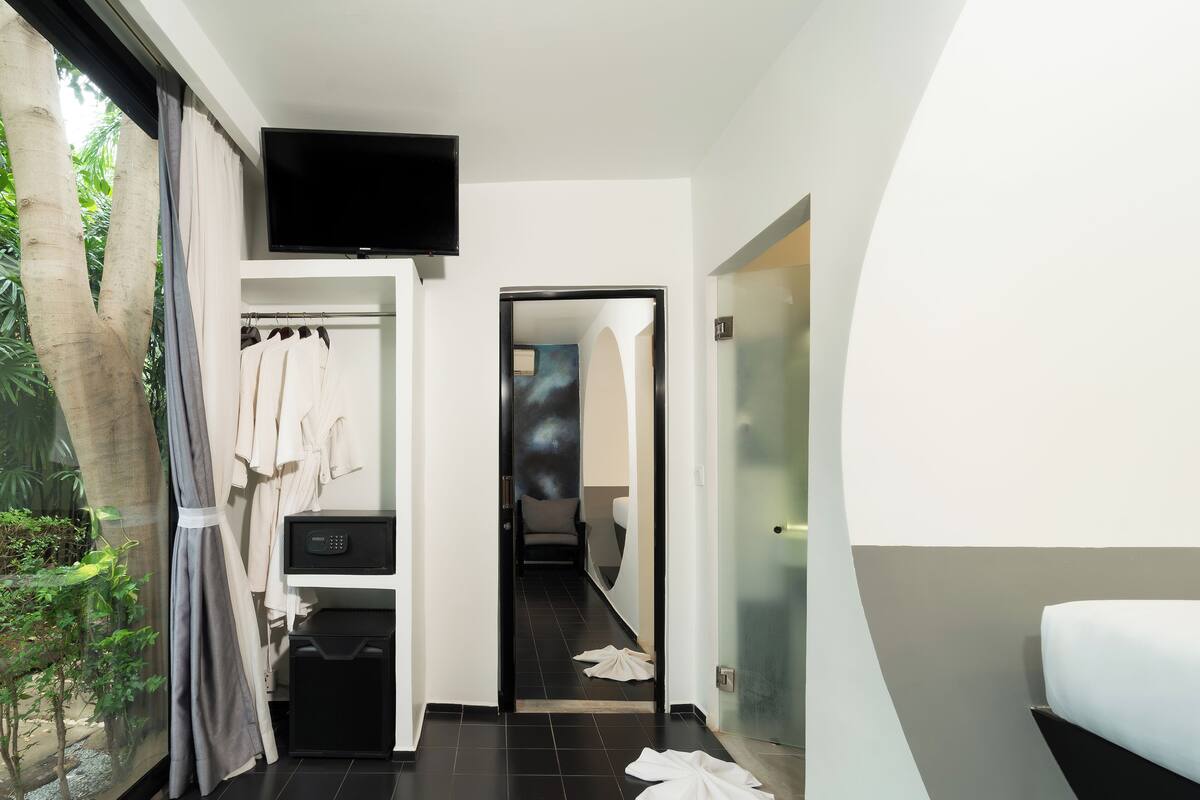 A modern hotel room with a large window, a flat-screen TV, a walk-in closet, and a bathroom with a glass door.