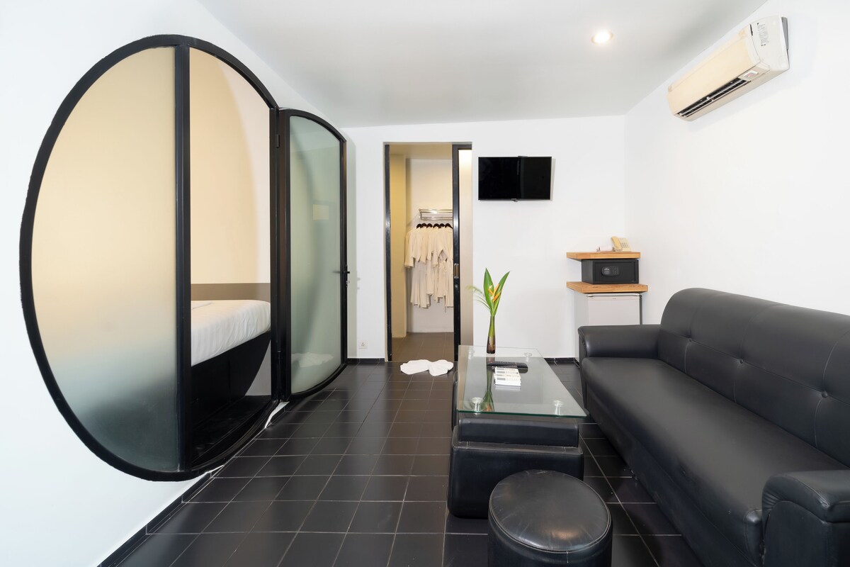 A modern hotel room with a glass partition, a black leather sofa, a glass coffee table, and a flat-screen TV mounted on the wall.