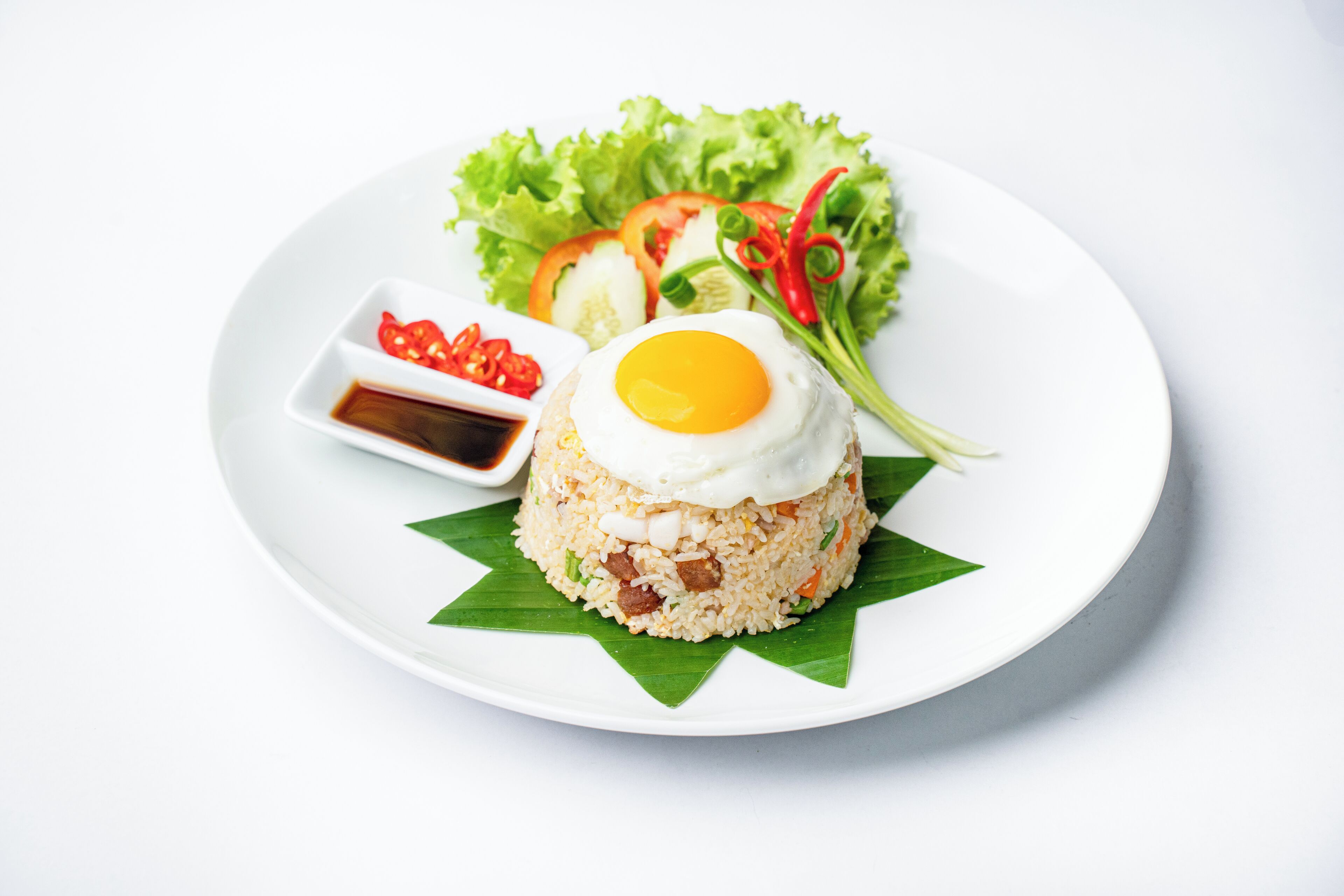A plate of fried rice with a fried egg on top, served with a side of vegetables and a small bowl of sauce.