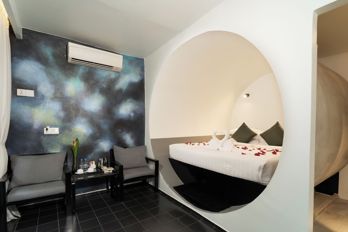 A modern hotel room with a unique curved bed, a galaxy-themed wall, and a cozy seating area with a small table and a chair.