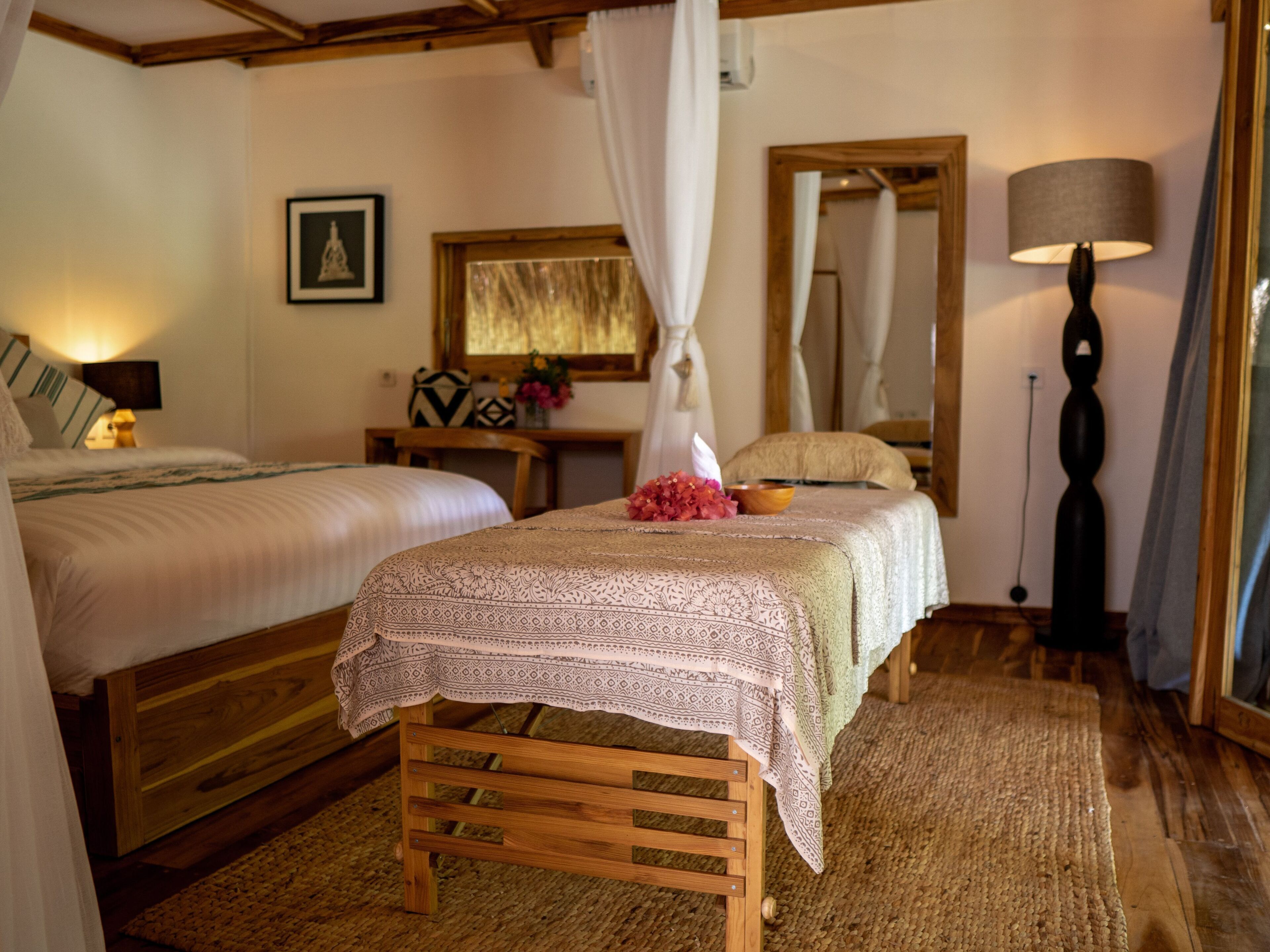 A cozy bedroom with two beds, a wooden bench, a bedside table with a lamp, and a mirror on the wall.