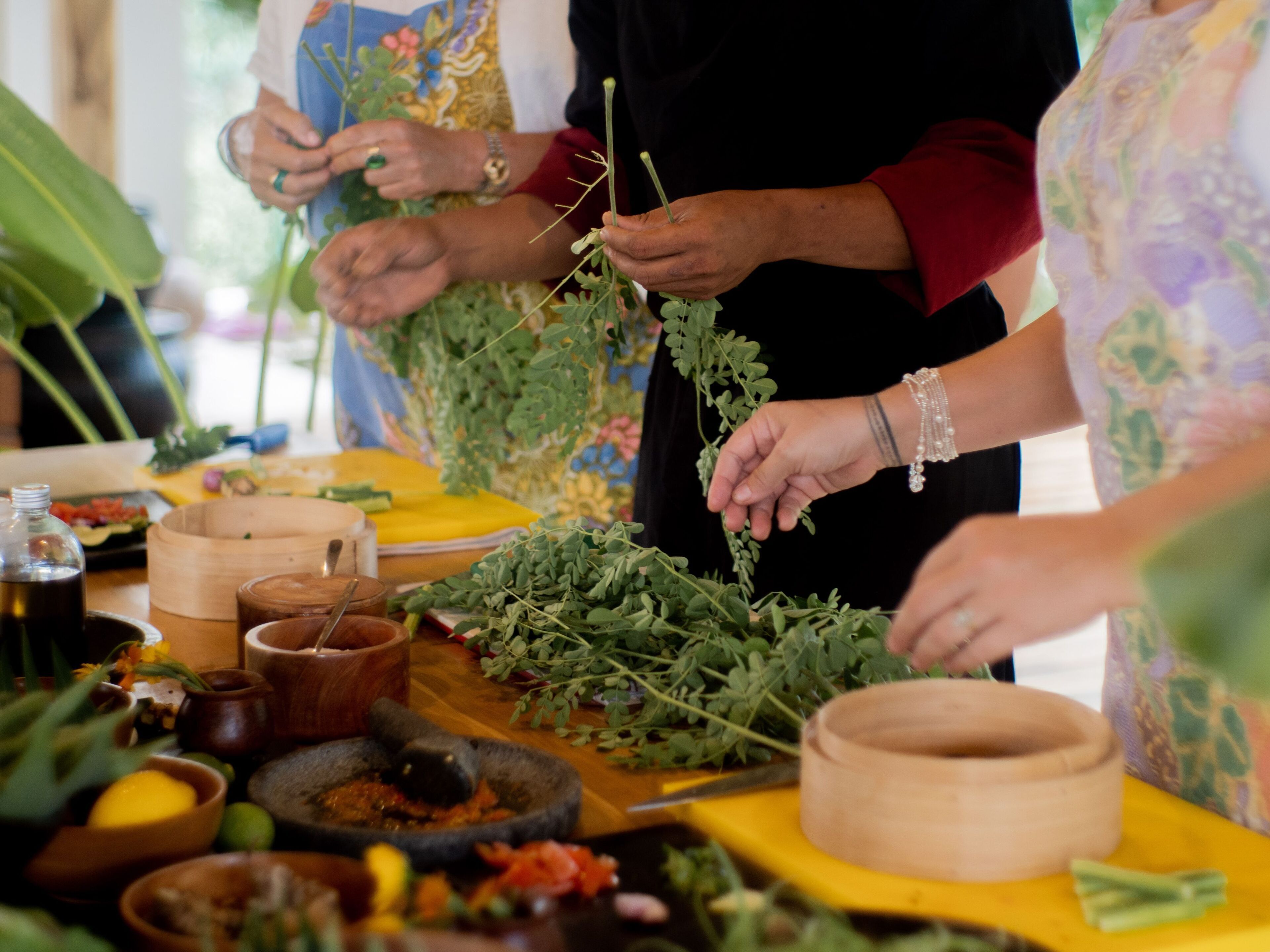 Un groupe de personnes prépare des plats à l’aide d’herbes fraîches et de divers ustensiles de cuisine, le tout sur une table en bois.