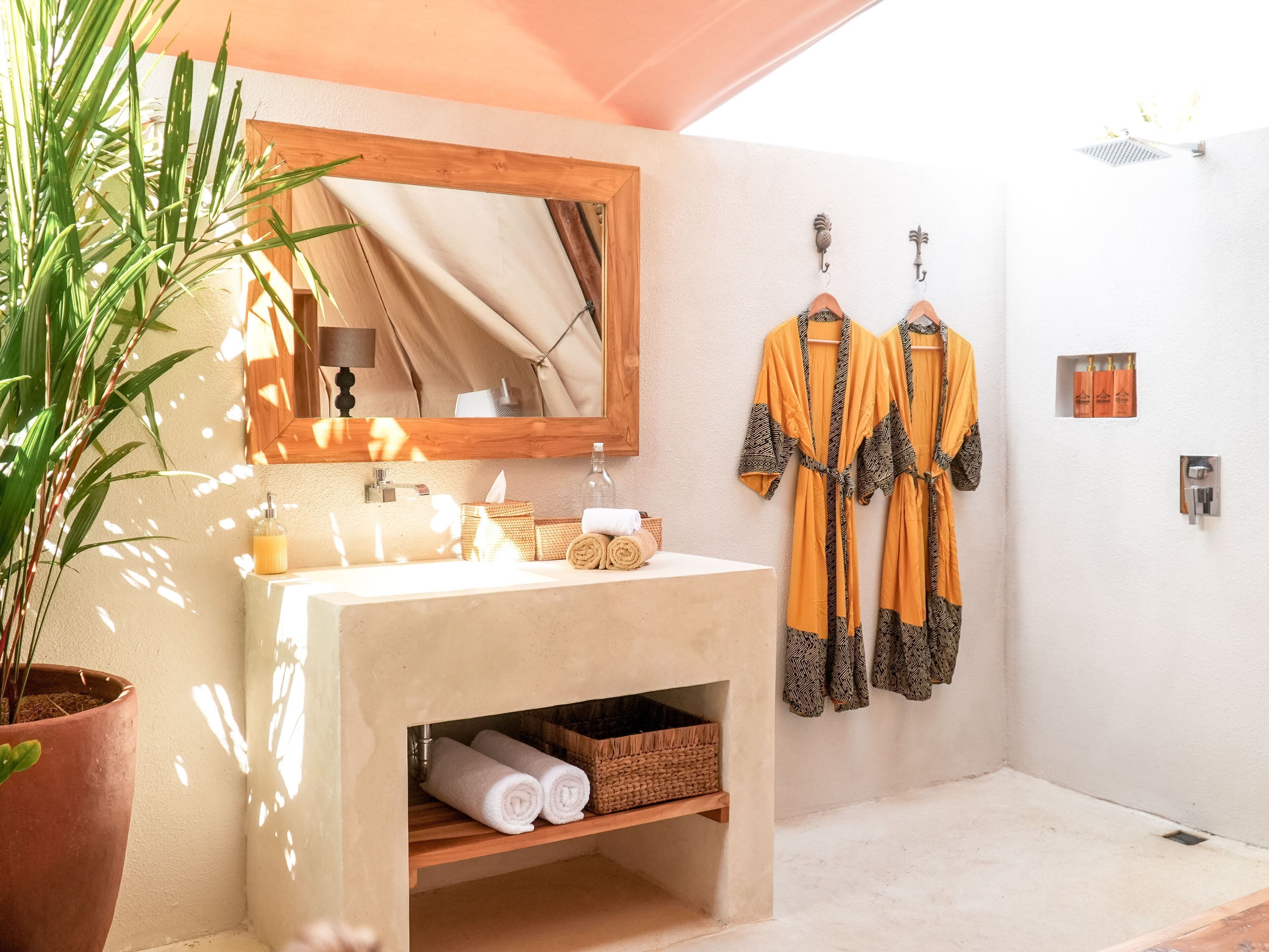 Een badkamer met een houten wastafelmeubel, een spiegel, een potplant, handdoeken en badjassen aan haken.