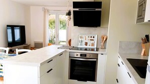 A modern kitchen with white cabinetry, a central island, a built-in oven, and a flat-screen TV. - The P'tit Jard near the beach (Jard-sur-Mer)