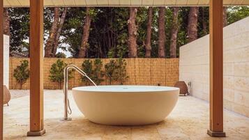 A large, round, white bathtub under a wooden pergola with a bamboo fence in the background.