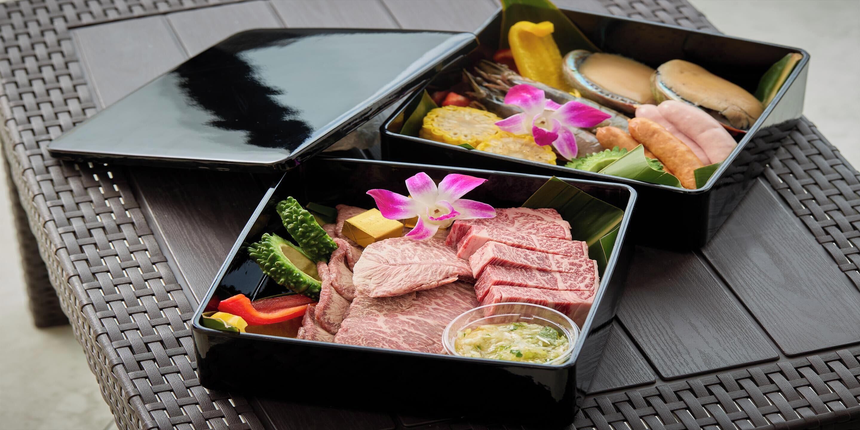Two black metal containers with food items, including meat, vegetables, and condiments, placed on a woven table.