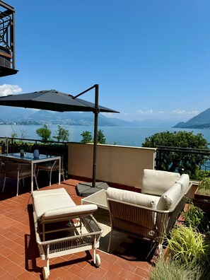 A terrace with outdoor furniture, a table, and an umbrella, overlooking a lake and mountains.