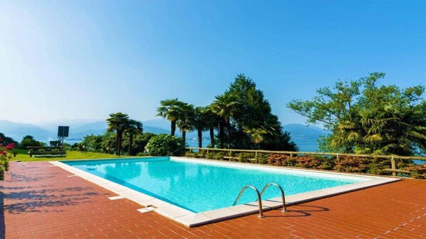 A rectangular swimming pool with a wooden deck and metal handrails, surrounded by lush greenery and palm trees, with a clear view of distant mountains and a body of water.