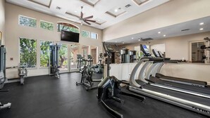 Une salle de sport spacieuse, équipée de divers appareils d’exercice, dotée de grandes fenêtres et d’un ventilateur au plafond.