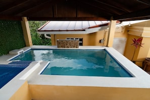 Una piscina rectangular con borde blanco, pared de piedra y estructura de techo metálica.