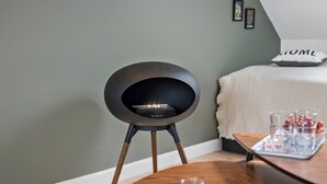 Une chambre moderne avec une cheminée ronde et noire posée sur un socle en bois, un lit recouvert d’une couverture blanche et un plateau avec des verres et des en-cas sur une table en bois.
