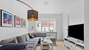 Un salon moderne avec un canapé gris, une table basse blanche et un téléviseur posé sur un meuble blanc. Des œuvres d’art encadrées ornent le mur, et une fenêtre offre une vue sur les bâtiments à l’extérieur.