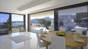 Un comedor moderno con una vista panorámica al océano, una mesa redonda de madera dispuesta para cuatro personas y un amplio ventanal con vistas al mar y a las montañas.
