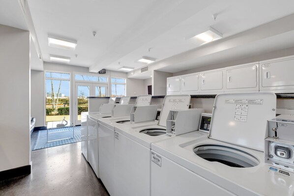A laundry facility with multiple washing machines and dryers, a blue mat on the floor, and a view of the outside through large windows.