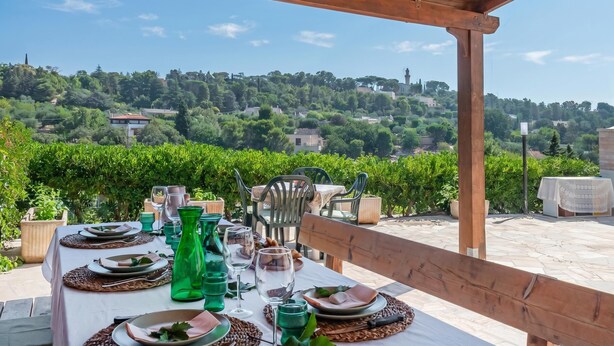 Un espacio de comedor al aire libre con una mesa preparada para cuatro personas, con vistas a un viñedo y una colina.