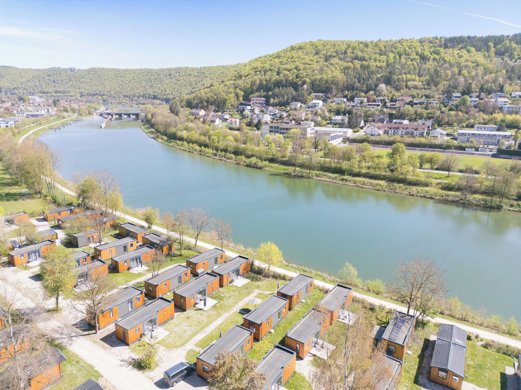 Vue sur un quartier résidentiel avec des maisons modernes, depuis la rive d’une rivière, et un pont visible au loin.