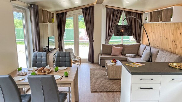 Ein modernes Wohnzimmer mit einer Couch, einem Esstisch und Stühlen. Ein Fernseher steht auf einem Schrank, eine Lampe ist vorhanden, und es gibt ein Fenster mit Vorhängen.