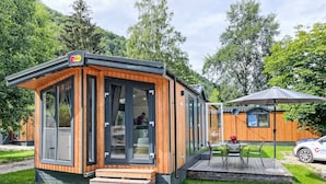 Eine moderne, kompakte Holzhütte mit großen Fenstern, inklusive einer kleinen Terrasse mit Sitzgelegenheit und Tisch.