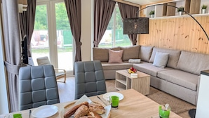 Ein modernes Wohnzimmer mit Sofa, Sesseln, einem Esstisch mit Brot und Getränken sowie einem Fernseher an der Wand.
