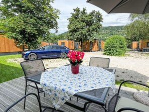Une terrasse avec une table et des chaises, une voiture bleue garée à proximité, et des cabanes en bois en arrière-plan.