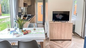 Une cuisine moderne avec des meubles en bois, une table à manger avec des chaises, un téléviseur posé sur un meuble en bois, et une vue sur l’extérieur.