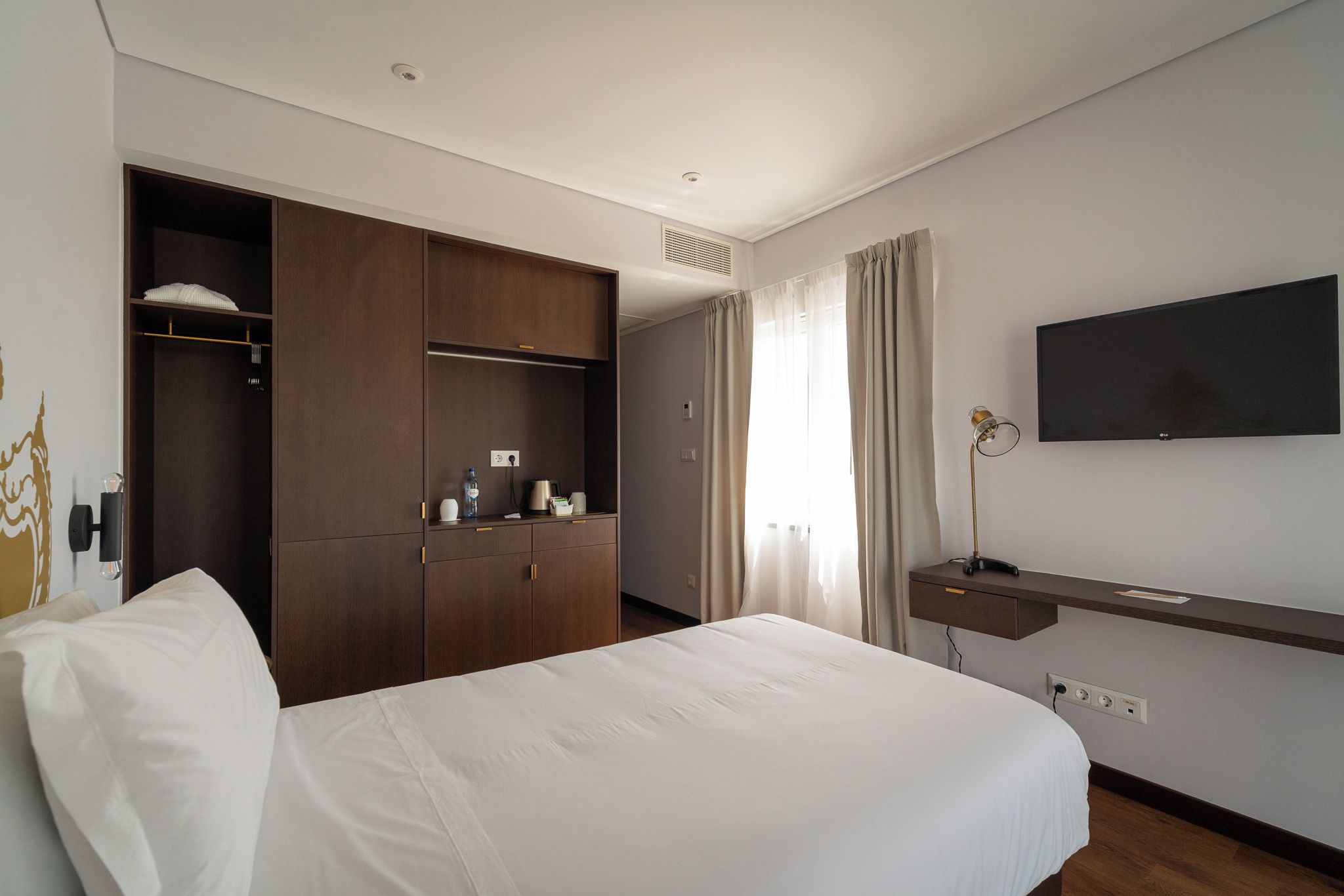 A hotel room with a bed, a wardrobe, a desk, a television, and a window with curtains.