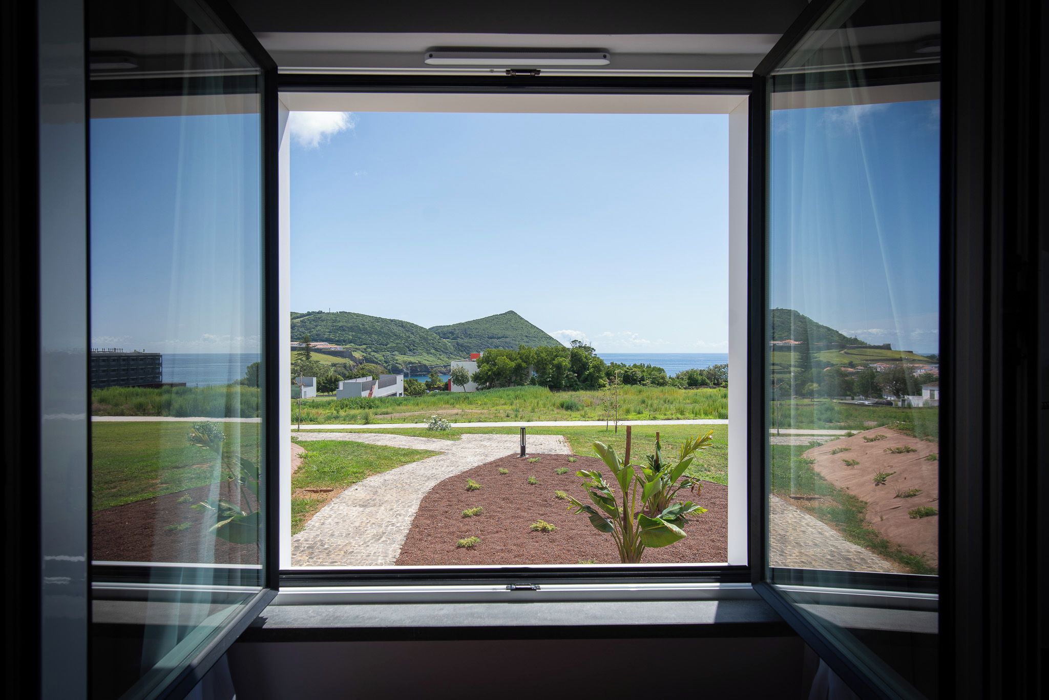 A window with a view of a landscaped area, a pathway, and distant hills.