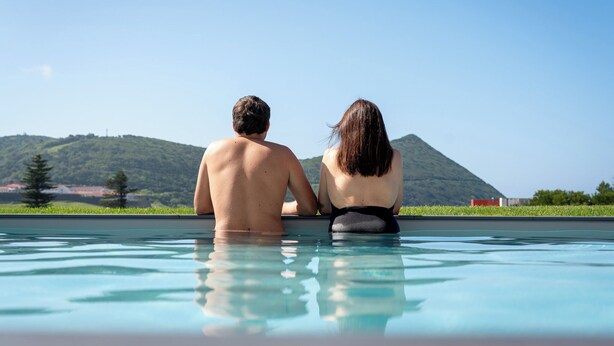 Two people sitting at the edge of a swimming pool with a view of a hill and trees in the background. - Palácio Santa Catarina (Angra do Heroismo)