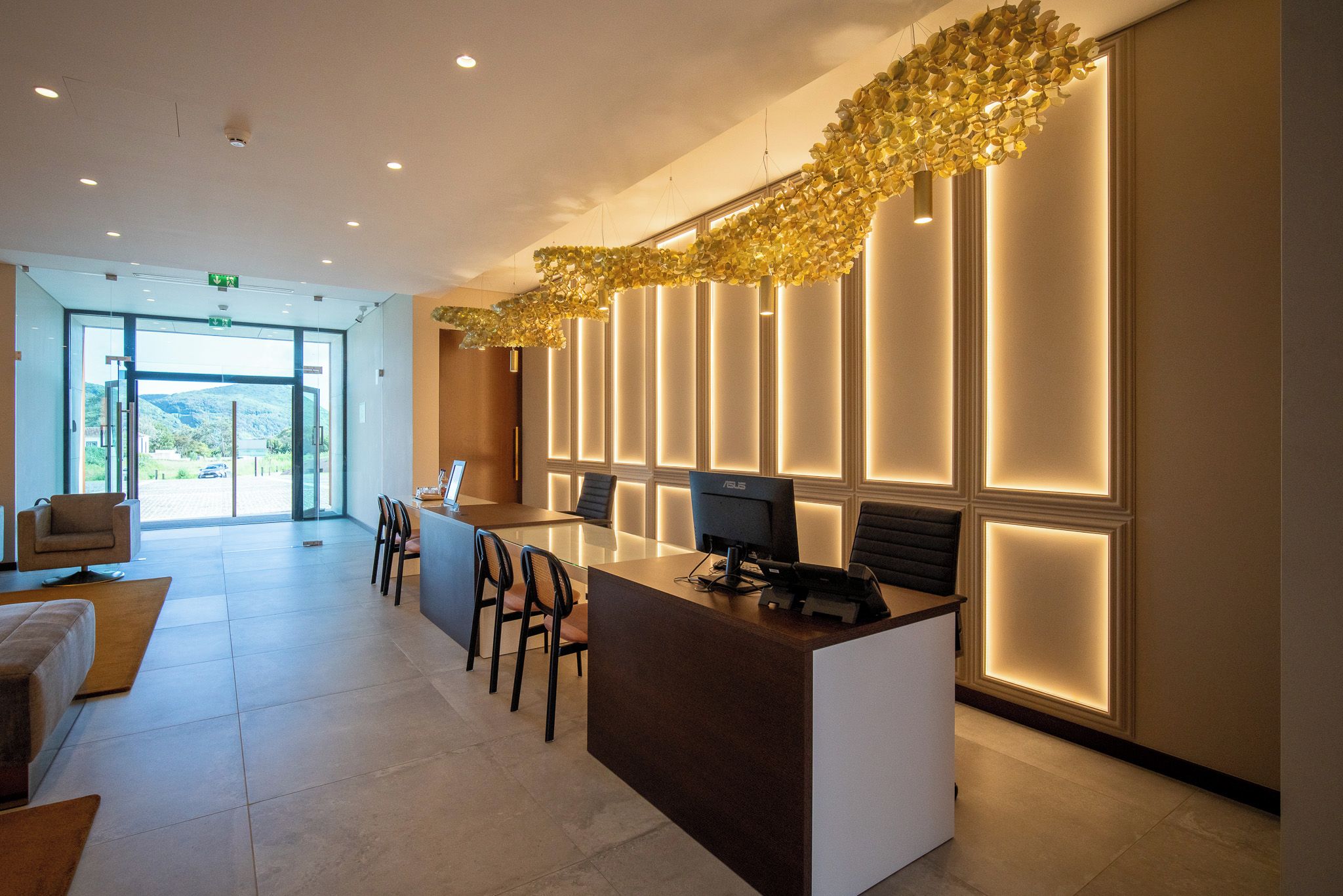 Een moderne receptie met een glazen deur naar buiten, een receptiebalie met stoelen en een decoratieve lamp erboven.