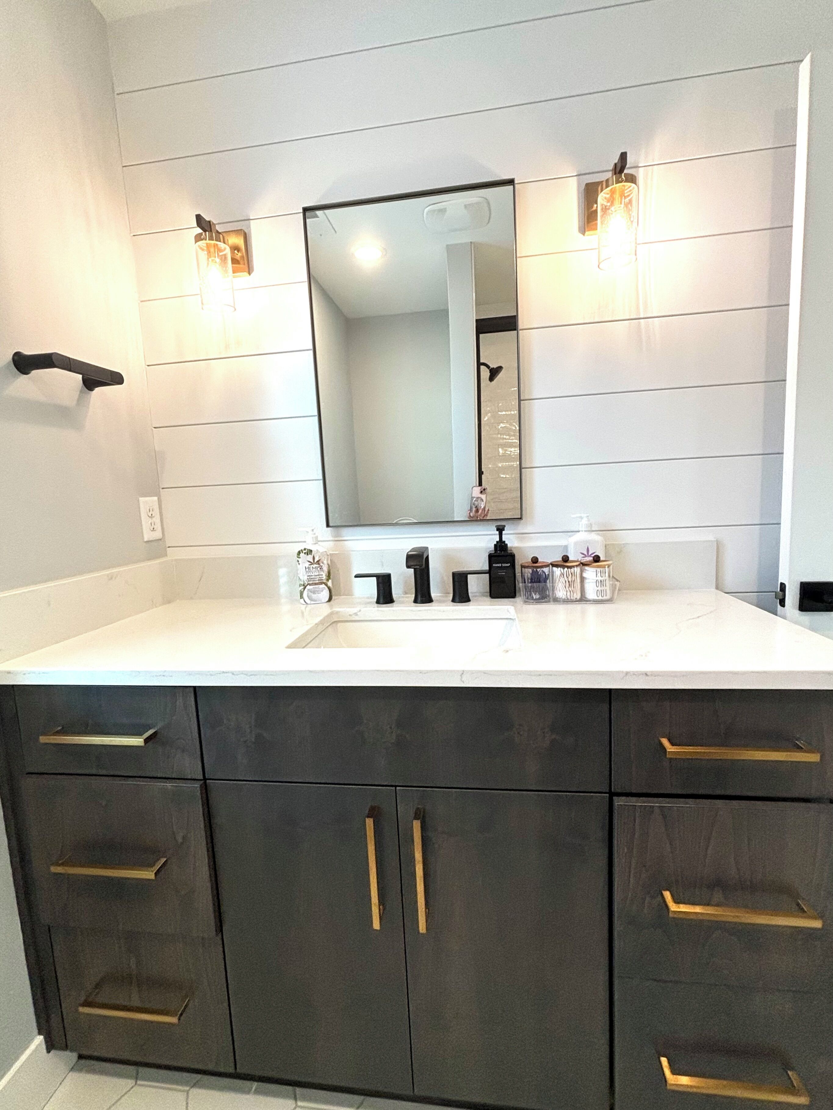 A bathroom with a white vanity, dark cabinets, and a large mirror. There are two wall-mounted lights and a faucet above the sink.