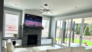 A modern living room with a stone fireplace, white leather sofas, a marble coffee table, and a large TV mounted above the fireplace. - Charming house with Beautiful Sunset View (Dallas)