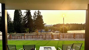A patio with a table and chairs, overlooking a fenced yard with a grassy area and trees.