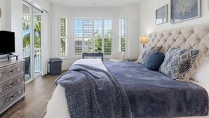 Un dormitorio con una cama grande, un ventilador de techo, televisión y vistas a palmeras a través de las ventanas.