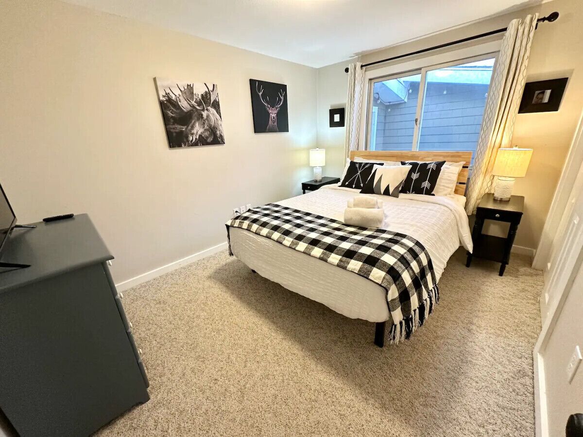 A bedroom with a bed, two wall-mounted lamps, a window with curtains, and two framed pictures on the wall.