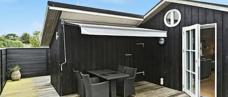 Una terraza de madera con una mesa y sillas, un porche cubierto y una puerta que da al interior.