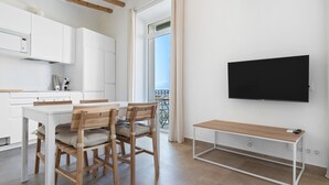 Une cuisine moderne avec des meubles blancs, une table à manger avec des chaises, un téléviseur fixé au mur et un balcon doté d’une balustrade.