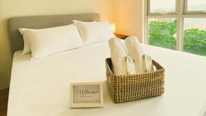 Un lit bien fait, avec du linge de lit blanc, un panier en osier rempli de serviettes et un panneau d’accueil.