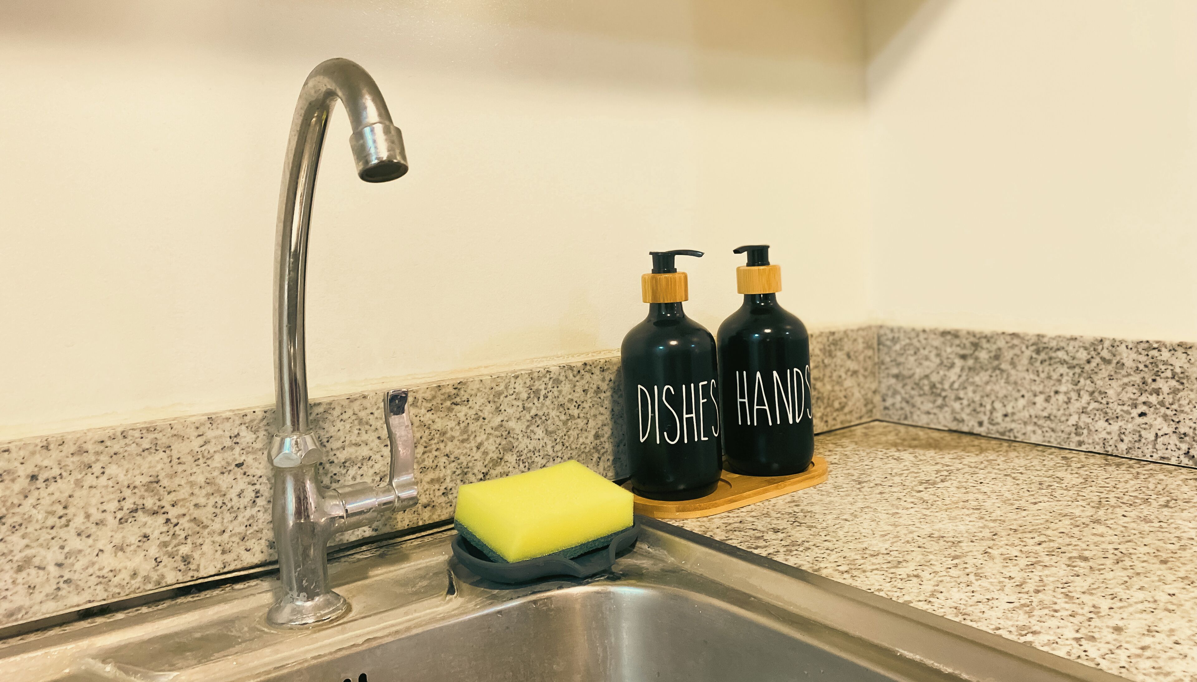 Un évier de cuisine avec un robinet, une éponge jaune et deux bouteilles noires étiquetées « VAISSELLE » et « MAINS » sur un plan de travail en granit.