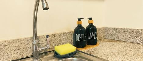 Un évier de cuisine avec un robinet, une éponge jaune et deux bouteilles noires étiquetées « VAISSELLE » et « MAINS » sur un plan de travail en granit.