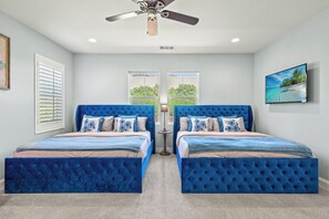 Ein Schlafzimmer mit zwei blau gepolsterten Betten, einem Deckenventilator und einem großen Fenster mit Blick auf Grünflächen.